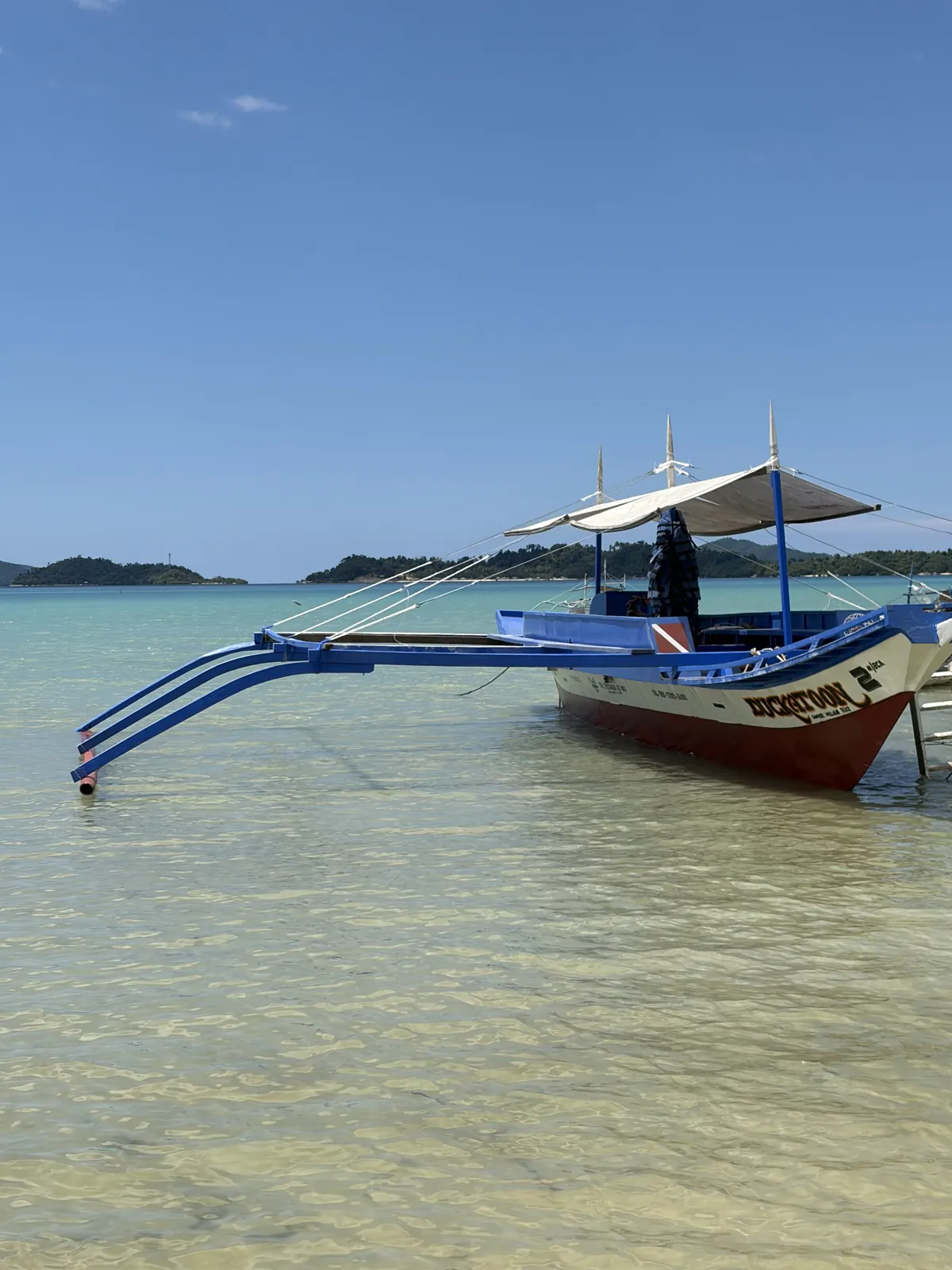 Bow and blue outriggers of Sunset Divers boat Dukatoon 2 in clear shallow water at Port Barton, Palawan